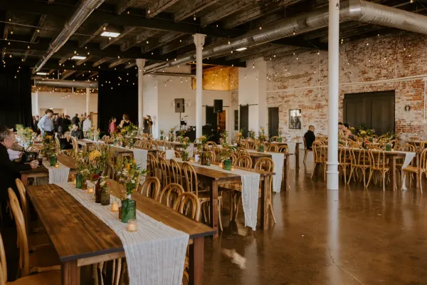 Wide interior shot of an affordable Indianapolis wedding venue showing exposed brick, original wood-beam ceiling, Edison string lights, and large industrial windows. Built-in venue character that does most of the decorating for couples on a budget.