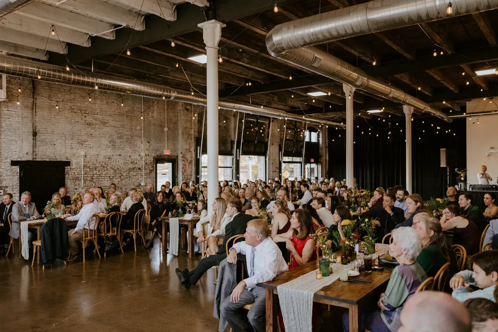 The Edison Room at 24 Shelby in downtown Indianapolis set for a corporate reception with round tables, ambient uplighting, and Edison string lights overhead against the original 1898 exposed brick walls.