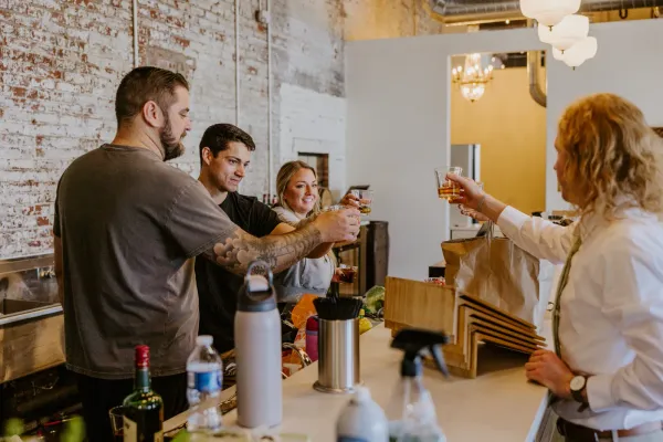 Guests raising cocktails in a toast at the Libations Lounge bar inside 24 Shelby, an affordable small party venue in downtown Indianapolis, with exposed brick and warm light behind them.