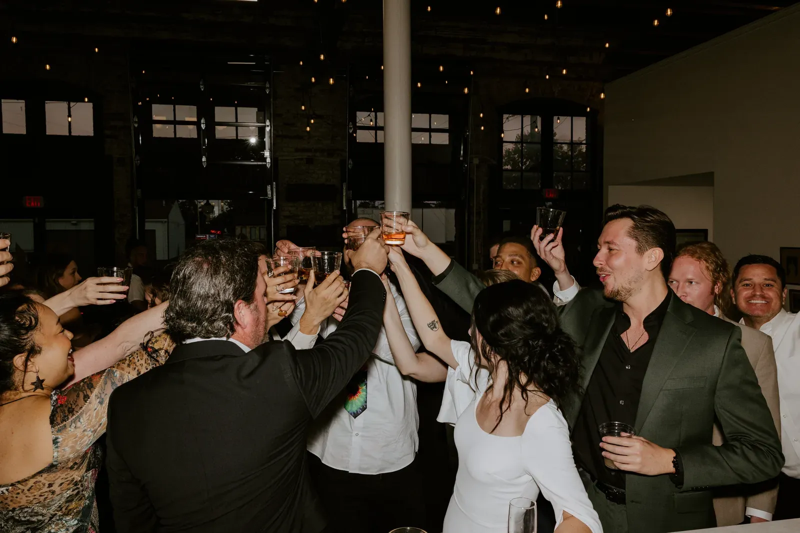 Guests raising a toast at a corporate event at 24 Shelby, a historic downtown Indianapolis event venue, under Edison string lights and original 1898 brick walls.