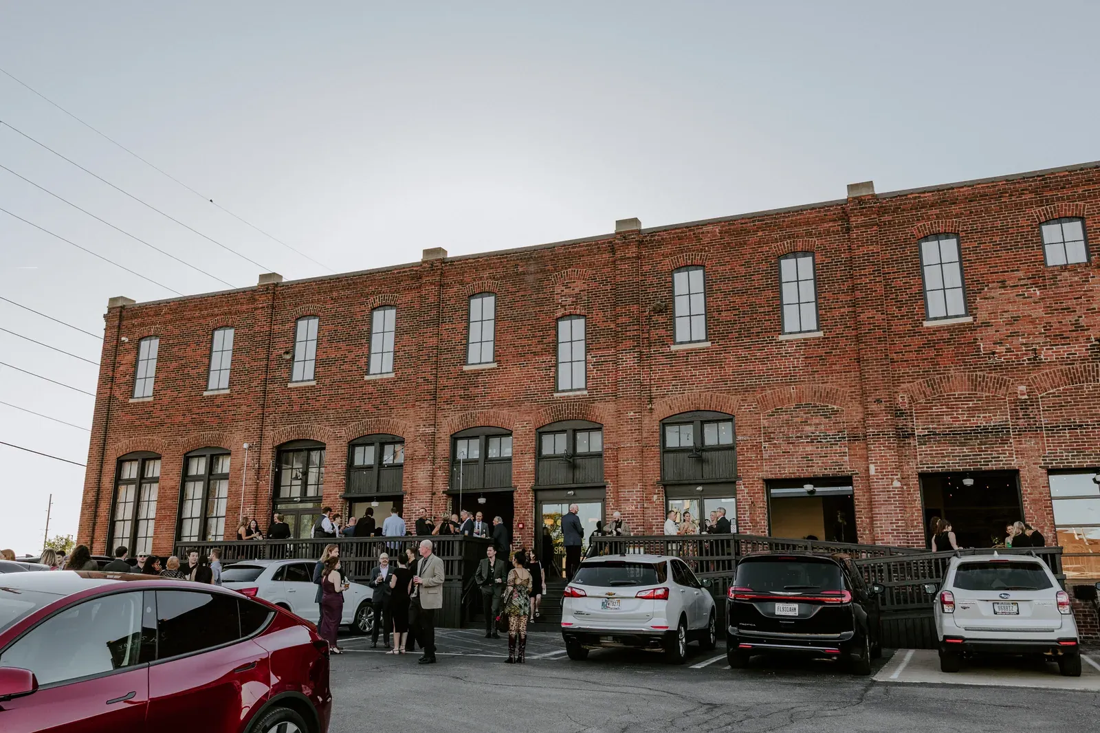 Wedding guests on the Prohibition Patio at 24 Shelby, an outdoor wedding venue in Indianapolis, during cocktail hour with the original 1898 brick facade and large industrial windows behind them.